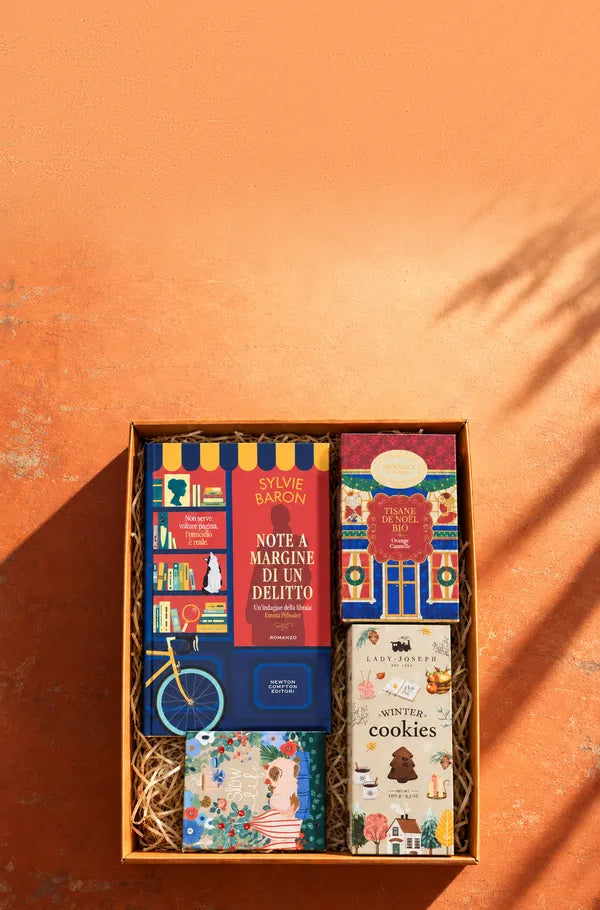 Una box Bookted aperta vista dall’alto con Note a margine di un delitto, una tisana natalizia all’arancia e cannella, i biscotti Winter cookies e un taccuino, su un muro arancio al sole
